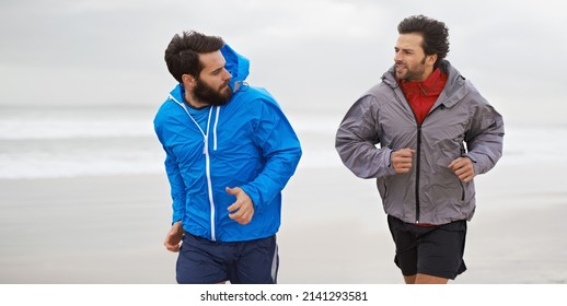 Taking A Morning Run On The Beach. Cropped Shot Of Two Young Men Jogging Together Along The Beach On An Overcast Morning.