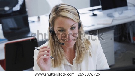 Taking notes mature support agent focusing on monitor at shared office desk, with headset and pen. Professional, collaboration, technology, modern, workplace, productivity, communication