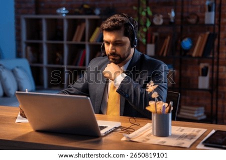 Taking notes in video call on laptop at office desk, Indian Asian businessman with headphones focused on virtual meeting, corporate executive engaged in remote work, digital communication