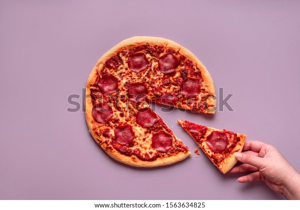 Taking one pizza slice. Woman hand\
grabbing a piece of pizza salami on a purple background. Above view\
of pepperoni pizza. Traditional italian\
dinner