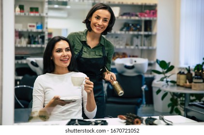 Taking A Rest. Close Up Photo Of A Sweet Woman Drinking Coffee And Looking In The Mirror While Talking With A Hairdresser In A Hair Salon.