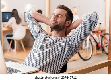 Taking Time For A Minute Break. Cheerful Young Man Holding Hands Behind Head And Keeping Eyes Closed While Sitting At His Working Place With His Colleagues Working In The Background 