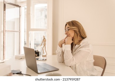 Talented adult caucasian woman sits in front of notebook and writes her new book in lightroom. Writer with blond hair is looking for inspiration. Work from home concept