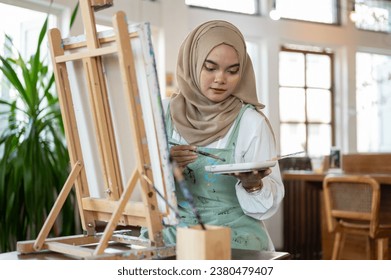 A talented Asian-Muslim female artist focuses on painting on canvas, mixing colors on a palette, working in her home studio. - Powered by Shutterstock