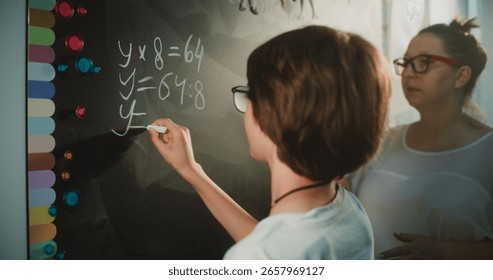 Talented Boy Demonstrating Math Knowledge: Close Up Shot of Primary School Student Solving Mathematical Equation Writing on the Board with Chalk in Classroom. Female Teacher Checking Correct Answers.