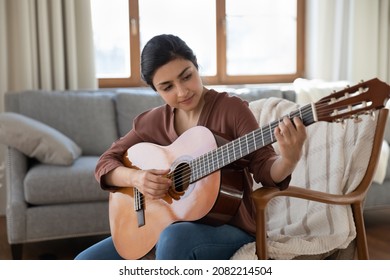 Talented concentrated young indian woman holding guitar in hands, practicing playing music learning chords, improving creative skills in modern living room, hobby leisure domestic activity concept.