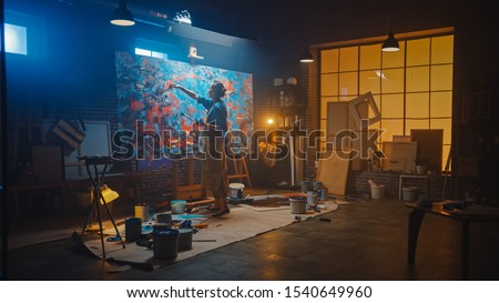 Talented Female Artist Working on a Modern Abstract Oil Painting, Gesturing with Broad Strokes Using Paint Brush. Dark Creative Studio Large Picture Stands on Easel Illuminated, Tools Everywhere