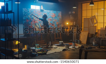 Talented Female Artist Works on Abstract Oil Painting, Using Paint Brush She Creates Modern Masterpiece. Dark and Messy Creative Studio where Large Canvas Stands on Easel Illuminated