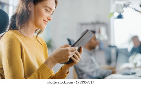 Talented Female Designer Sitting at Her Desk She's Holding and Using Touch Screen Digital Tablet Computer. Bright Office where Diverse Team of Young Professionals Work.