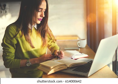 Talented female student of journalism creating article for homework using textbook and web sources searching information writing down ideas into copybook sitting in coworking space with laptop 