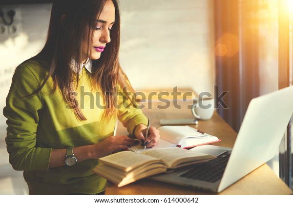 Talented female student of journalism creating article\
for homework using textbook and web sources searching information\
writing down ideas into copybook sitting in coworking space with\
laptop 