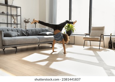 Talented gymnast kid doing upsidedown acrobatic handstand with splits in air, exercising on floor in living room. African American kid training body, sportive skills at home. Childhood, sport concept