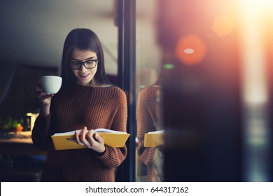 Talented pretty student of college reading interesting story for completing homework task,teen woman dressed stylish clothing enjoying leisure time with cup of coffee and good mood standing indoors