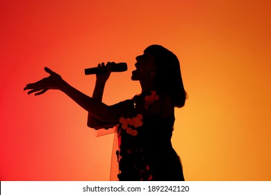 Talented. Silhouette of young female singer isolated on orange gradient studio background in neon light. Beautiful shadow in action, performing. Concept of human emotions, expression, ad, music, art.