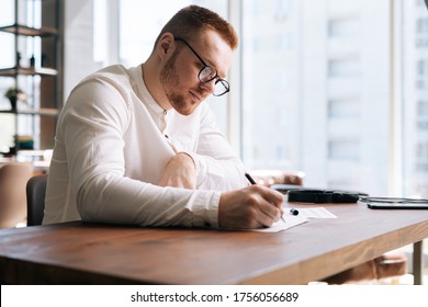 Talented young composer wearing stylish eyeglasses is writing musical notes with pen on paper sheet music sitting at the wooden desk in modern lighting room on background of large window in sunny day. - Powered by Shutterstock