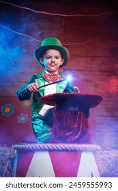 Tales of the Old Circus. A little boy with a painted mustache and a circus green costume plays magician with a magic wand and hat in the circus arena. 