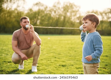 Talking by using paper cup telephone. Happy father with son are having fun on the field at summertime.