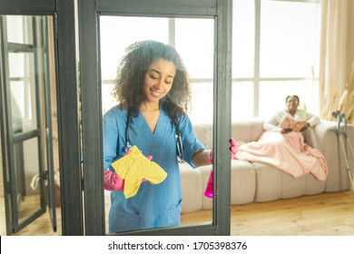 Talking And Cleaning. Pleasant Young Curly Nurse Talking To The Patient While Cleaning The Glass Door