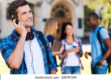 Talking With Friends. Handsome Young Man Talking On The Mobile Phone And Smiling While Standing Against University Building With His Friends Chatting In The Background