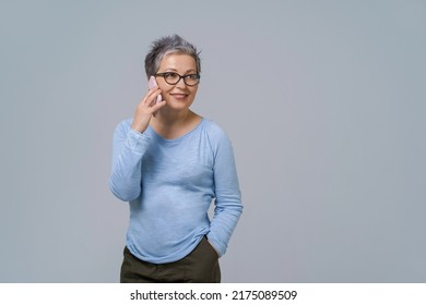 Talking On The Phone Mature Grey Hair Woman In Glasses Turn Sideways Holding Smartphone In Her Hand Wearing Blue Blouse And Black Skirt. Pretty Woman In Blue Shirt Isolated On White Background.