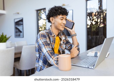 Talking on smartphone, teenage boy using laptop and smiling at home. Technology, communication, home office, multitasking, remote working, chatting