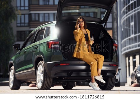 Talks by phone. Young fashionable woman in burgundy colored coat at daytime with her car.