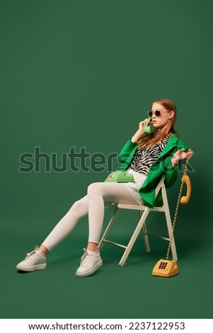 Talks. Creative portrait of young without emotions girl in green jacket sitting on chair with retro phone isolated over green background. Vivid style, beauty, queer, fashion concept. Copy space for ad