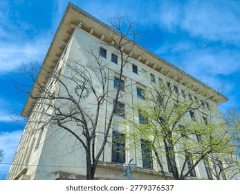 A tall building with multiple windows is seen against a blue sky. Trees bare few leaves surround the building. The setting appears to be a lively urban area during daylight.