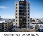 Tall building with a lot of windows and a snow covered roof. The building is in a city and is surrounded by other buildings