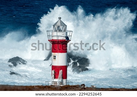 A tall circular lighthouse tower has horizontal red and white colors against a stormy sea. The lighthouse has steps, windows, a walkway, and metal walls. The building sits high on top of a rocky cliff