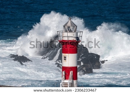 A tall circular lighthouse tower has horizontal red and white colors against a stormy sea. The lighthouse has steps, windows, a walkway, and metal walls. The building sits high on top of a rocky cliff