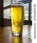 A tall clear pint beer glass filled with cold foam from a lager ale. The golden colored ale sits on the edge of a wooden patio table at a microbrewery. There