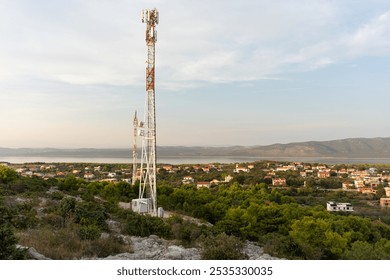 Tall communication towers on rocky hillside overlooking a small village and a scenic coastal landscape with distant mountains and calm water under clear sky. Modern technology and countryside scenery. - Powered by Shutterstock