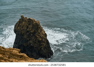 Tall, dark rock formation is being pounded by waves from the rough ocean on an overcast day - Powered by Shutterstock