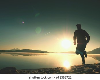 Tall fit man running fast by the sea on the beach. Powerful runner training outdoor in summer morning. Hot Sun is rising up over water level