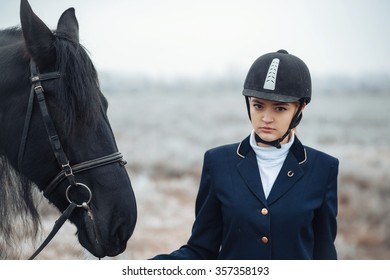 A Tall Girl With Long Blond Hair In A Jockey Outfit With A Beautiful Black Horse In An Empty Snow-covered Field In Winter