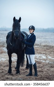 A Tall Girl With Long Blond Hair In A Jockey Outfit With A Beautiful Black Horse In An Empty Snow-covered Field In Winter