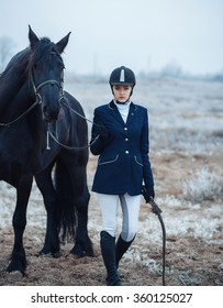 A Tall Girl With Long Blond Hair In A Jockey Outfit With A Beautiful Black Horse In An Empty Snow-covered Field In Winter