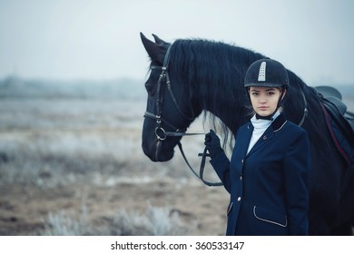 A Tall Girl With Long Blond Hair In A Jockey Outfit With A Beautiful Black Horse In An Empty Snow-covered Field In Winter