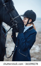 A Tall Girl With Long Blond Hair In A Jockey Outfit With A Beautiful Black Horse In An Empty Snow-covered Field In Winter