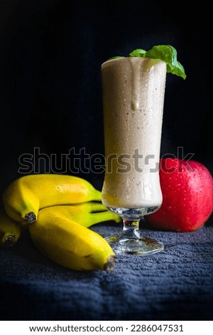 A tall glass of banana shake garnished with a mint leaf, served on a fabric napkin with bananas and apples, all styled in darkfood style. Perfect photo for a  cafe menu, or a culinary blog.