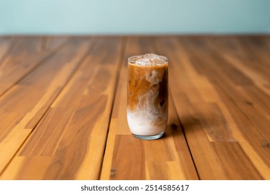 A tall glass of iced coffee with milk swirls, sitting on a wooden surface against a soft blue background. - Powered by Shutterstock