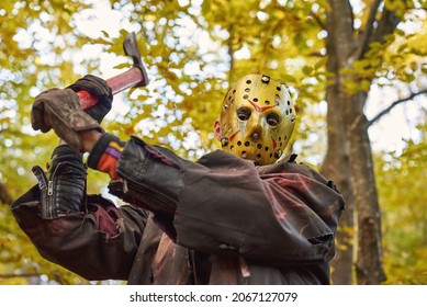 Tall guy with halloween mask holds wooden axe in the forest.