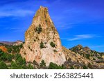 A TALL JAGGED ROCK STRUCTURE WITH SEVERAL TREES GROWING ON IT WITH GREEN TREES IN THE FORWGROUND WITH A DEEP BLUES SKY IN THE GARDEN OF THE GODS IN COLORADO SPRINGS COLORADO