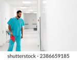 A tall male doctor with a beard walks along the hospital corridor with a folder of documents and enters his office