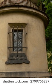 A tall, narrow window set in a rounded plaster tower wall, fitted with vertical and horizontal metal bars and a decorative stone sill beneath a small tiled
