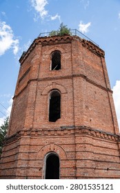 A tall octagonal red brick tower with multiple arched window openings and decorative brick cornices stands abandoned under a bright blue sky with scattered