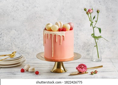 Tall pink cake decorated with macaroons, raspberries and chocolate balls on golden cake stand over white background with flowers and berries. Side view, copy space - Shutterstock ID 1822426775