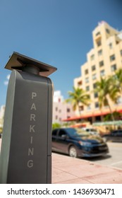 Tall and rectangular electronic and solar powered public parking meter with vertical computer generated parking illustration letters against a bokeh street and building background