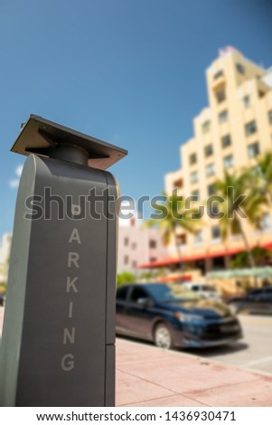 Tall and rectangular electronic and solar powered public parking meter with vertical computer generated parking illustration letters against a bokeh street and building background